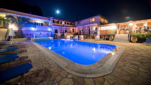 A pool area with lounge chairs and a stone pathway leading to a building with multiple levels and balconies. - Arianna Studios And Apartments (Corfu)