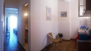 A narrow hallway leading to a bathroom with a sink and a chair. There are framed pictures on the wall and a window with curtains. - Arianna Studios And Apartments (Corfu)