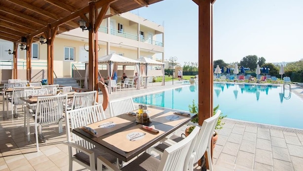 Ein überdachter Außenbereich mit Tischen und Stühlen, mit Blick auf einen Pool und ein Gebäude mit Balkonen.