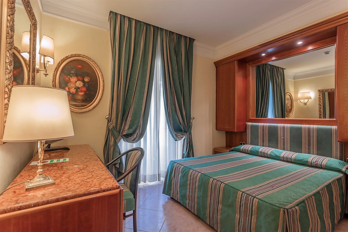 A hotel room with a bed, a desk, a chair, a lamp, a mirror, and curtains.