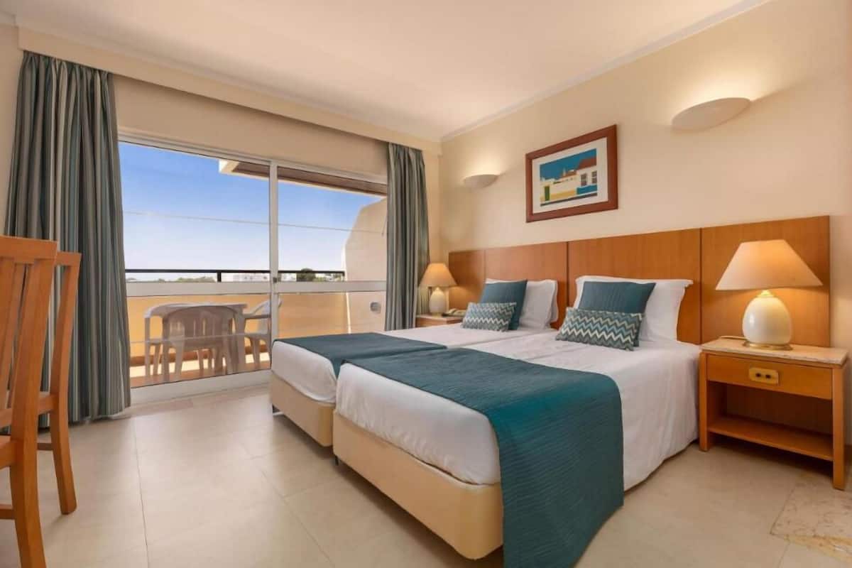 A hotel room with two beds, a balcony with a view, and a painting on the wall.