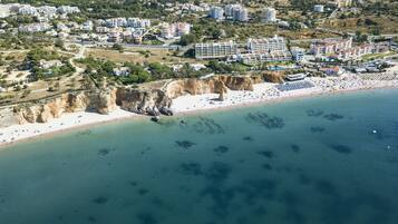 Vista aerea di una zona costiera con spiaggia sabbiosa, scogliere rocciose e un fitto gruppo di edifici sullo sfondo.