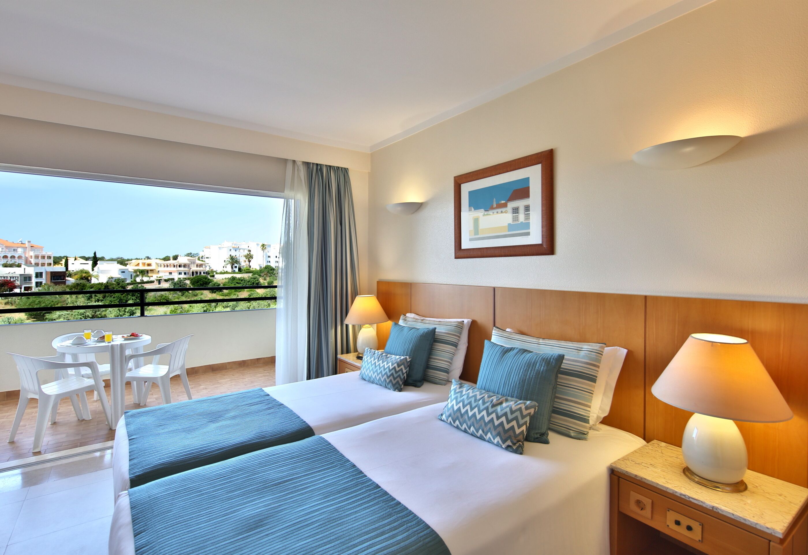 Habitación de hotel con dos camas, un balcón con mesa y sillas, y vistas a edificios.