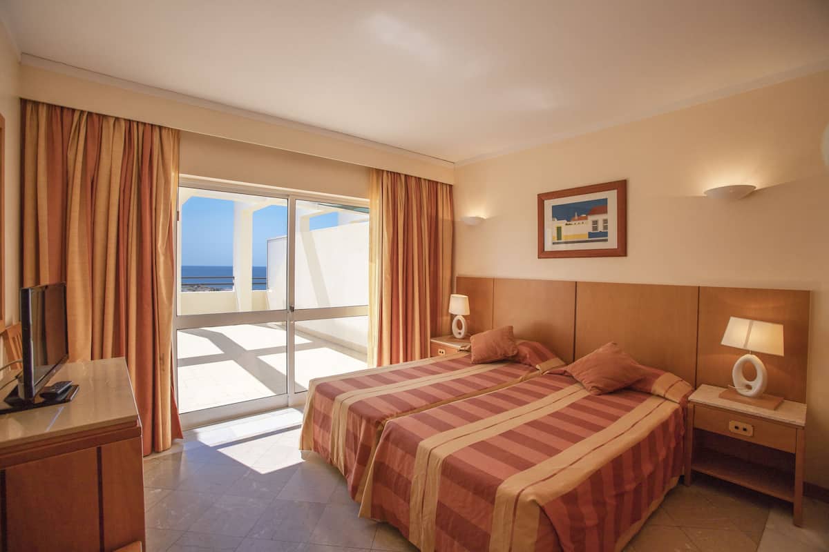 A hotel room with a large bed, a TV, a desk, and a view of the sea through a sliding door.