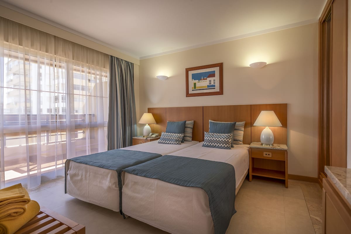 A hotel room with two beds, a large window with sheer curtains, a painting on the wall, and a bedside table with lamps.