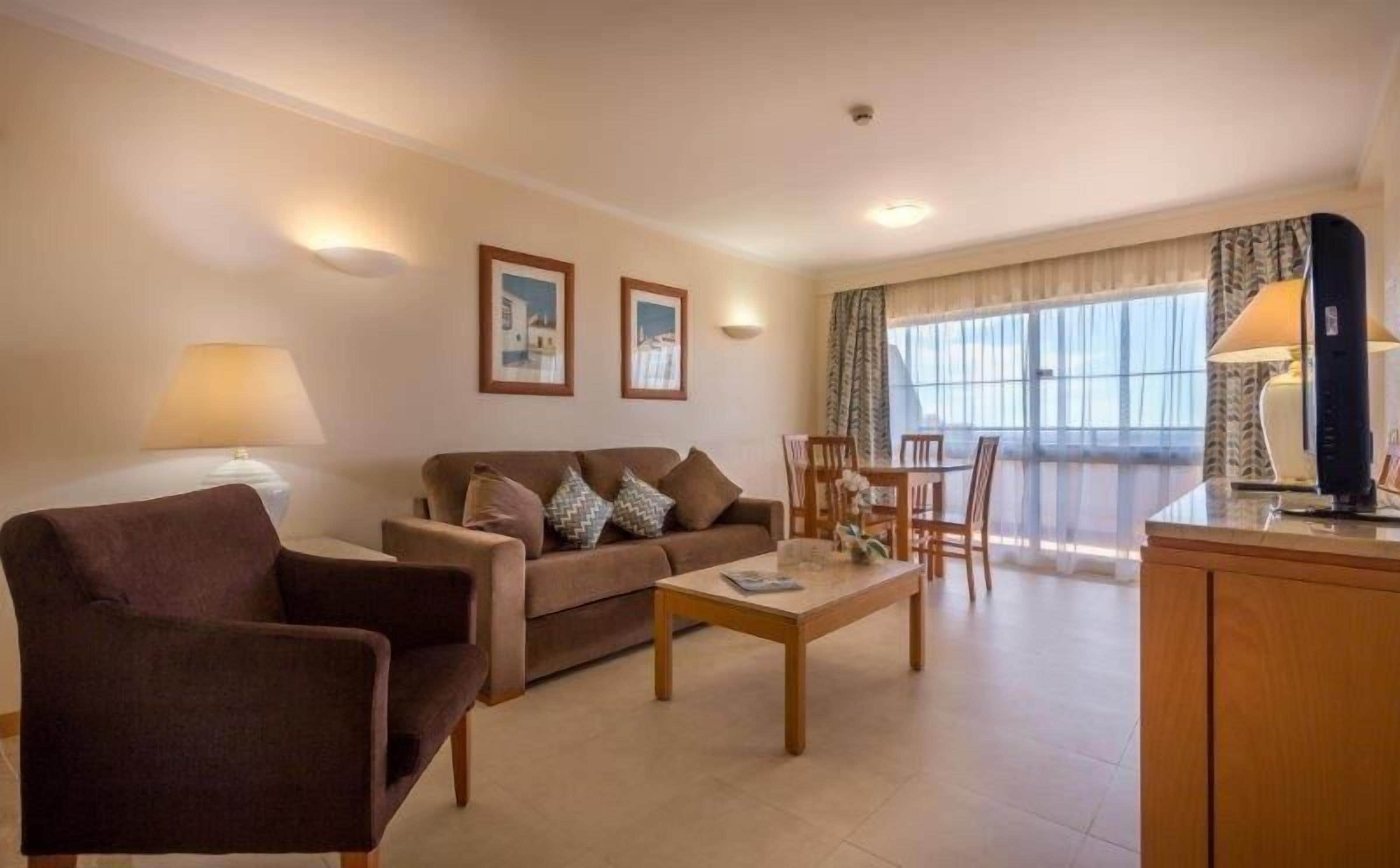 Uma sala de estar com sofá, poltrona, mesa de centro e área de refeições. Há duas quadros nas paredes, uma luminária e uma janela com cortinas.