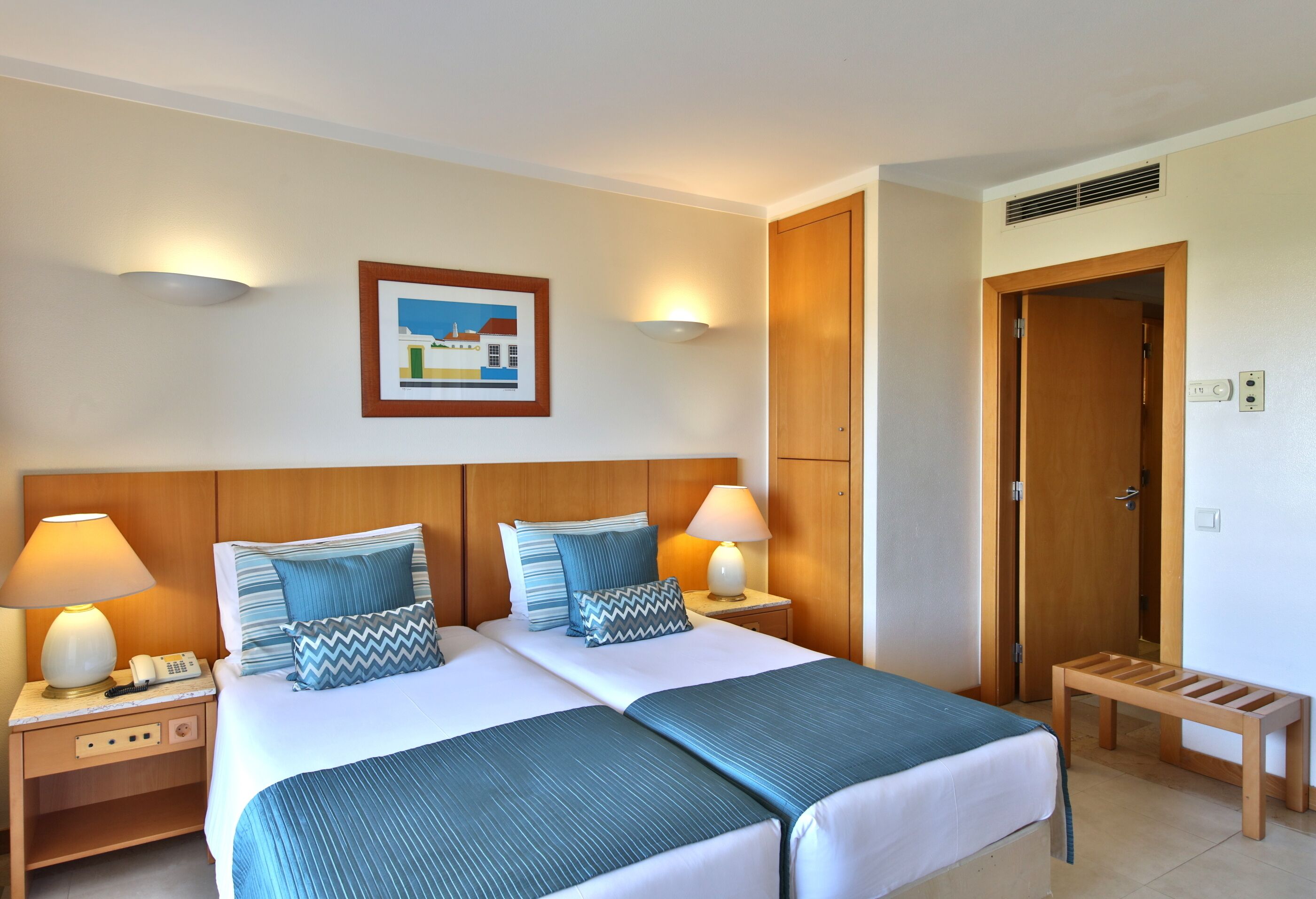 A hotel room with a double bed, two bedside tables with lamps, a wooden headboard, a framed picture on the wall, and a door to the right.