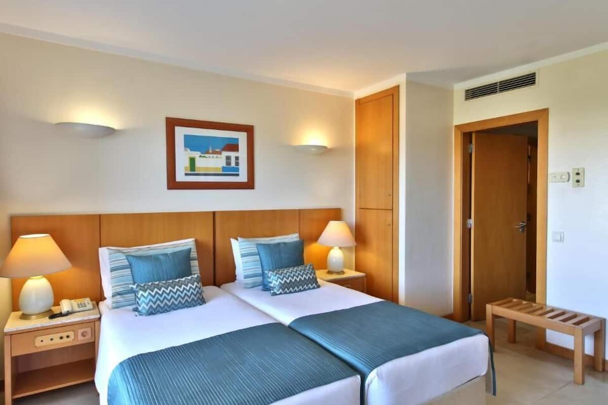 A hotel room with a double bed, two bedside tables with lamps, a wooden headboard, a framed picture on the wall, and a wooden bench.
