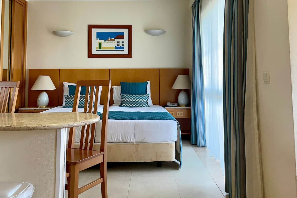 A hotel room with a bed, a desk with a chair, a trash bin, and a large window with curtains.