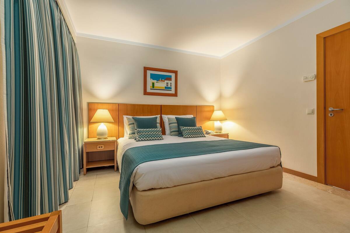 A neatly arranged bedroom with a large bed, two bedside tables with lamps, a wooden headboard, a framed picture on the wall, and a wooden door.
