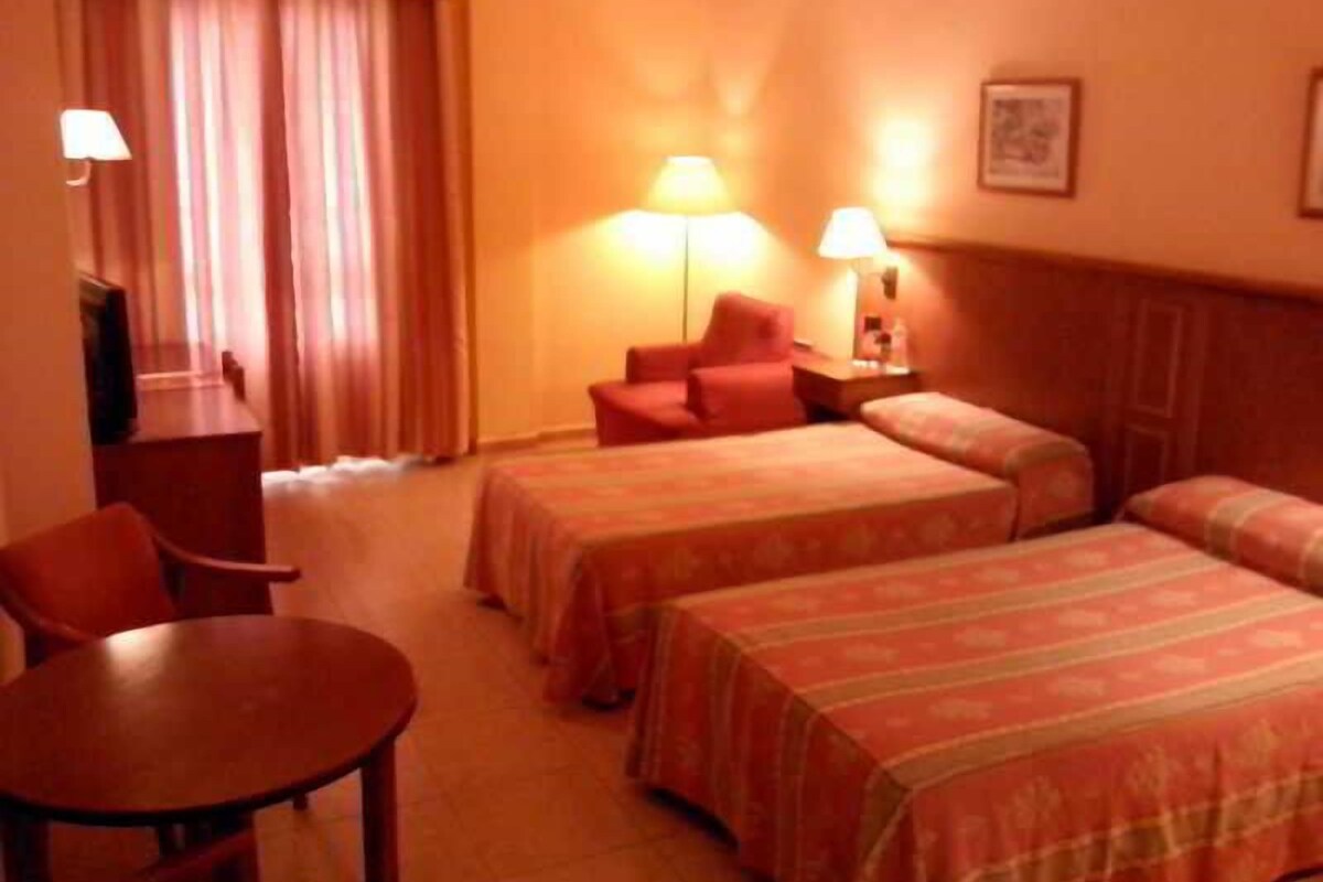 A hotel room with two beds, a desk, a chair, and two lamps.