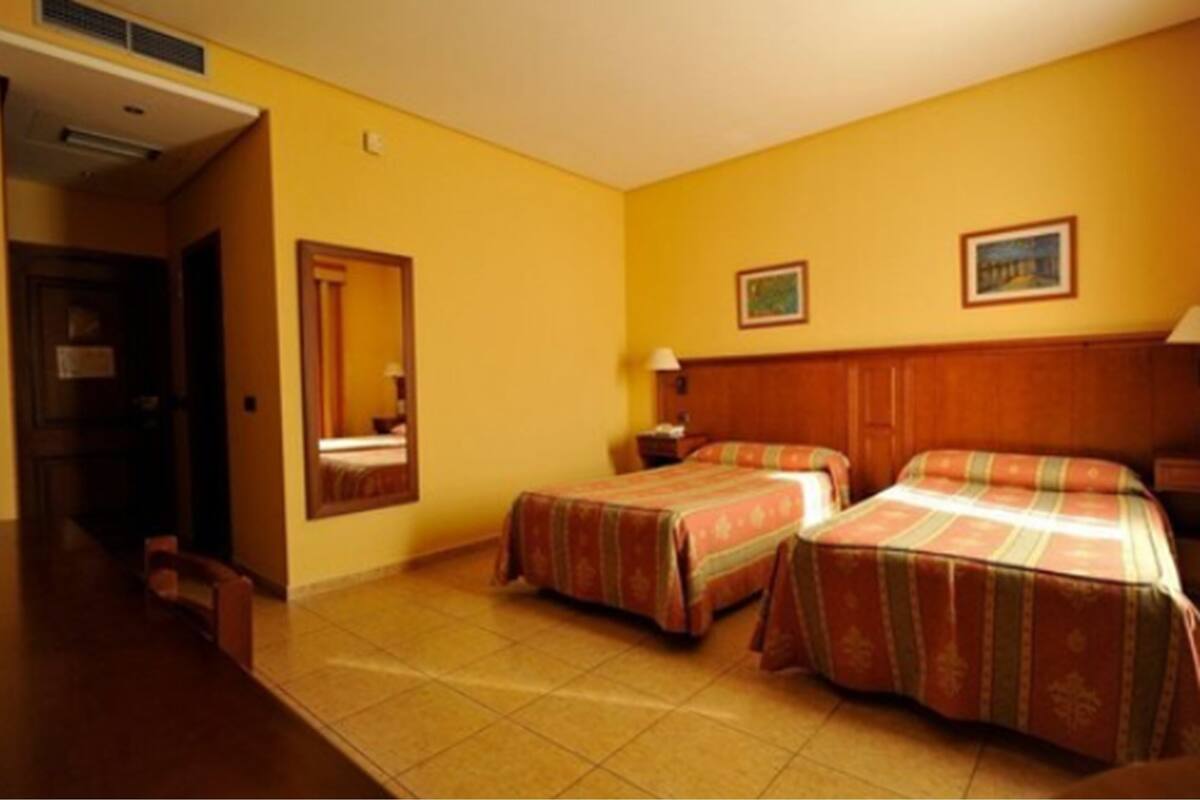 Une chambre d’hôtel avec deux lits, une tête de lit en bois, une table de chevet, un miroir et une porte donnant sur une autre pièce.
