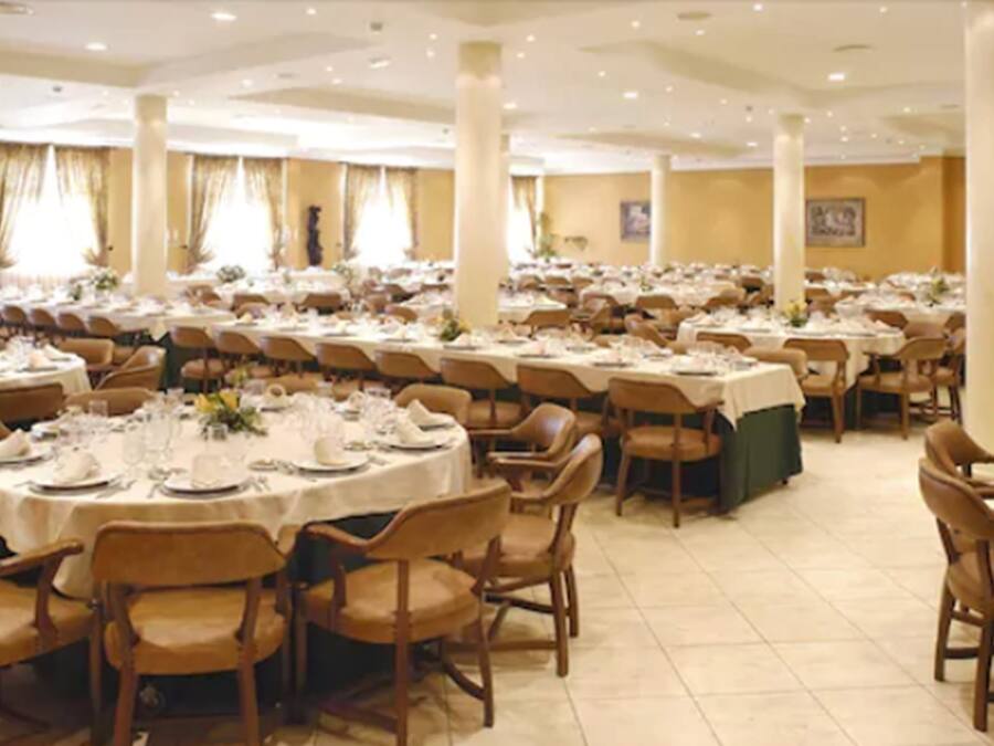 Um amplo salão de banquetes com mesas redondas dispostas para refeições, rodeado por cadeiras de madeira.