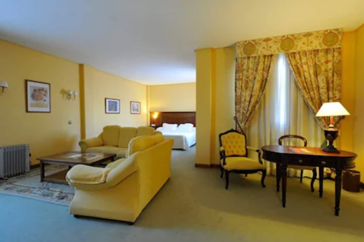 Une chambre d’hôtel avec un canapé jaune, une table à manger avec des chaises, une lampe et un lit en arrière-plan.