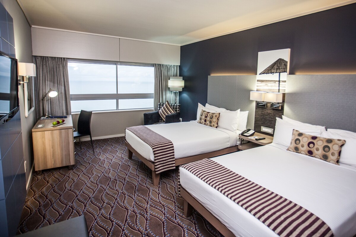 Habitación de hotel con dos camas, un escritorio, una silla, una lámpara y vistas al mar.