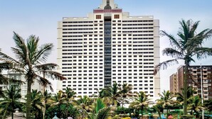 A tall hotel with a central glass elevator shaft, surrounded by palm trees and a pool. - Garden Court Marine Parade (Durban)