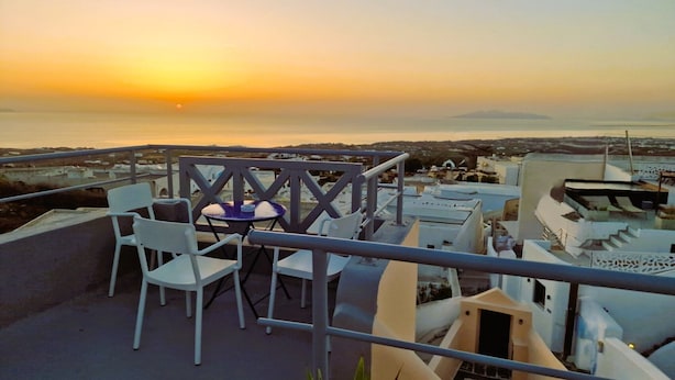 Una terraza con sillas blancas y una pequeña mesa redonda, con vistas a un paisaje urbano y al mar al atardecer.