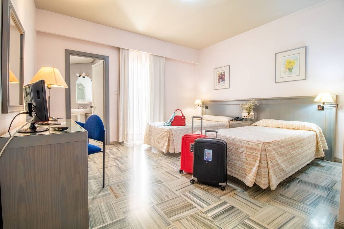 A hotel room with a desk, two beds, a mirror, a window with curtains, and a bathroom visible through an open door.