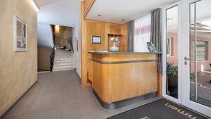 Zona de recepción de un hotel con un mostrador de madera, una puerta de cristal que da a un balcón y una escalera al fondo.