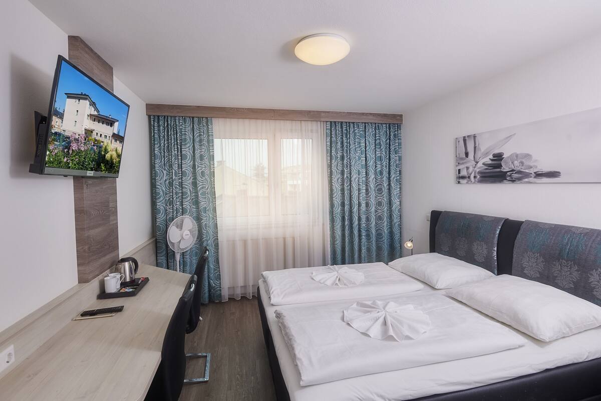 Una habitación de hotel con cama, escritorio, televisor, ventilador y ventana con cortinas.