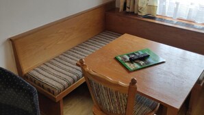 A wooden bench with a cushioned seat, a wooden table with a remote control, and a window with bamboo curtains. - Pension Eder (Saalbach-Hinterglemm)