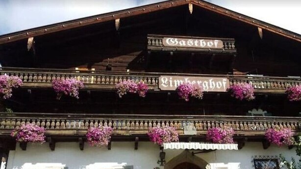 Ein traditionelles Holzgebäude mit Balkonen, die mit rosa Blumen geschmückt sind, und Schilder mit der Aufschrift „Gasthof“ und „Limburghof“.