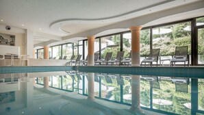 En stor inomhuspool med väggar av genomskinligt glas, omgiven av solstolar och med utsikt över grönområden utanför.