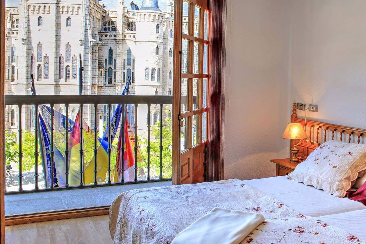 A bedroom with a bed, a nightstand, a lamp, and a view of a castle through a window.