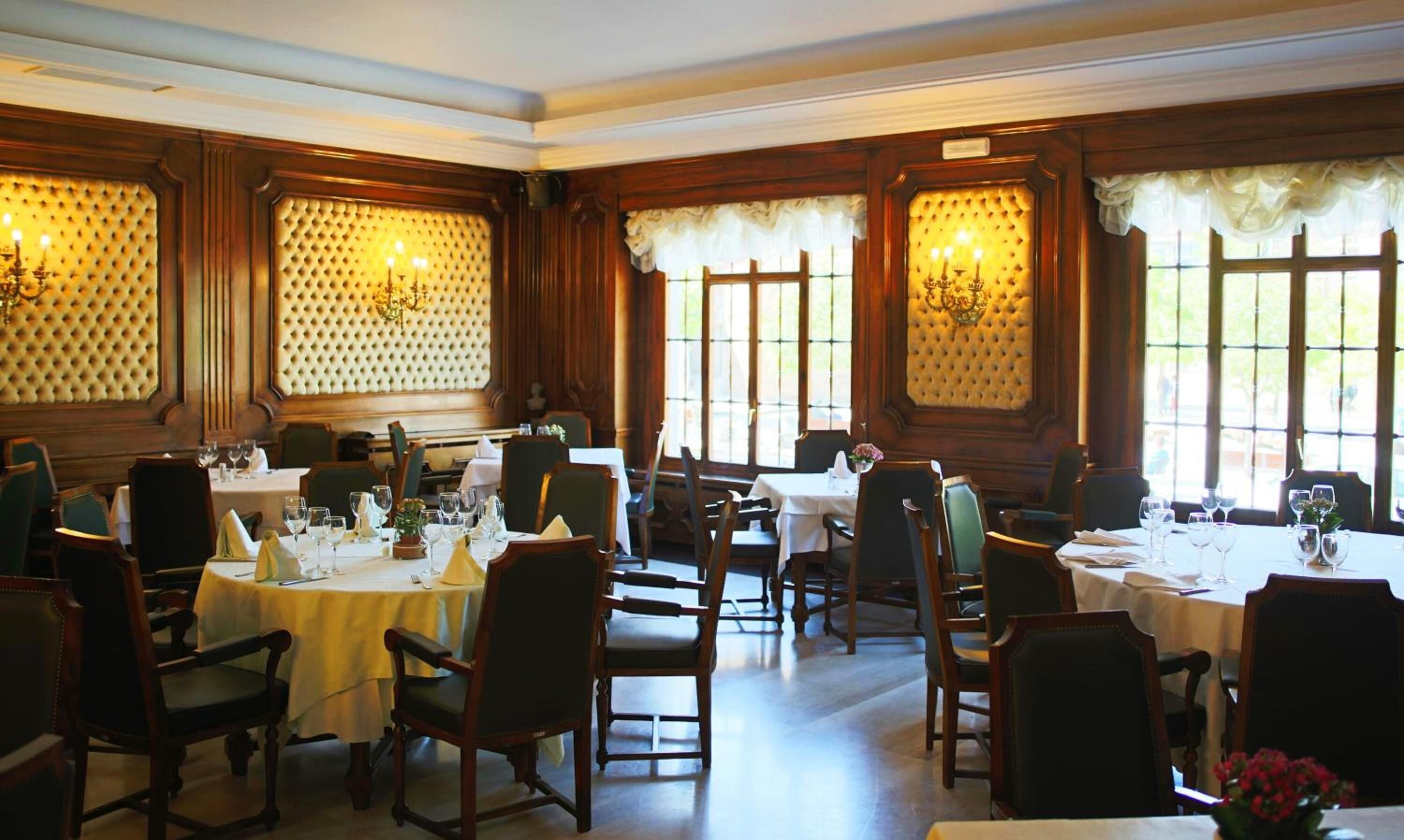 A dining area with multiple tables set for service, large windows with curtains, and wall paneling.