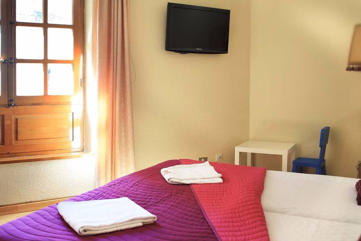 A hotel room with a bed, a television on the wall, a window with curtains, a bedside table, and a lamp.