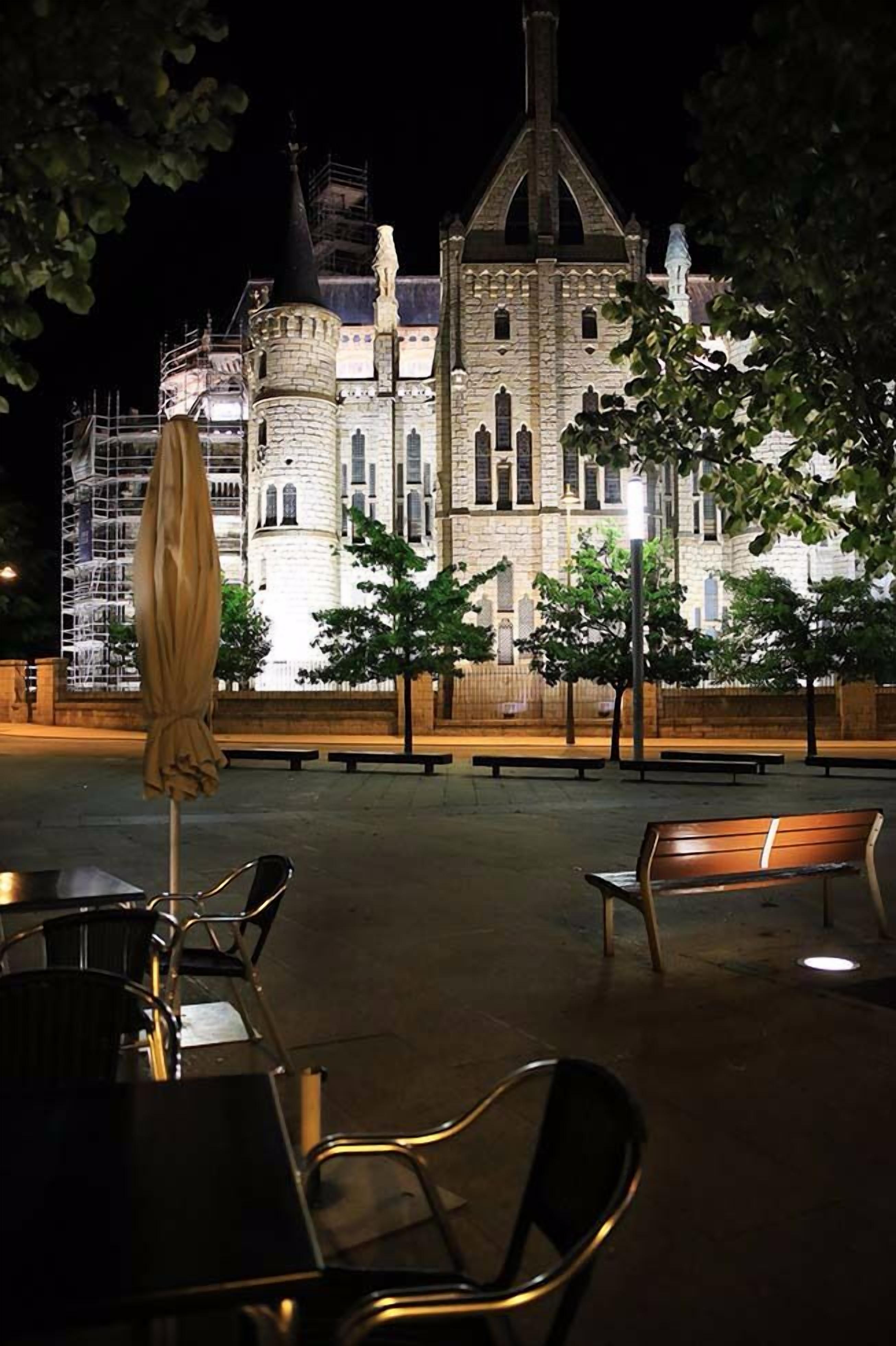 Un espacio al aire libre con mesas, sillas y un banco, con un edificio grande al fondo.