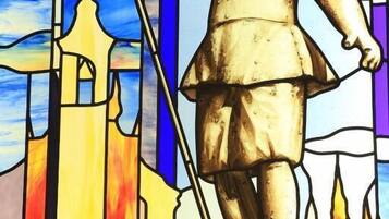 Una ventana de vitral con una figura sosteniendo un báculo, frente a un cielo colorido y un fondo arquitectónico.