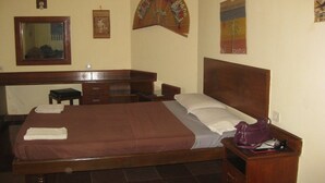 A hotel room with a bed, wooden headboard, a desk with a chair, a small stool, and a nightstand with a bag and a book. - Mobor Beach Resort (GOA)