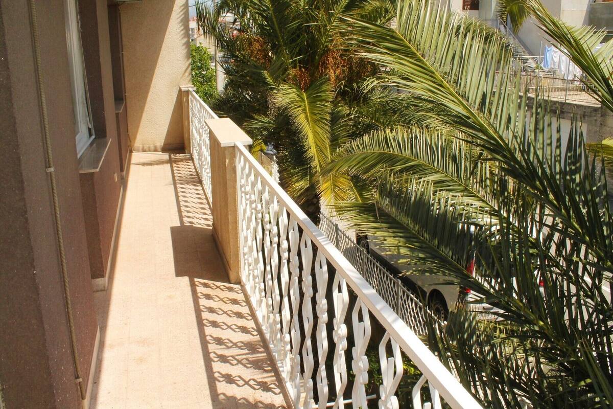 Un balcon avec une balustrade blanche, des palmiers et, au fond, un bâtiment avec un toit recouvert de tuiles.