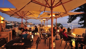 Un área de comedor al aire libre con mesas y sillas bajo sombrillas, con vistas a un paisaje urbano y al mar.