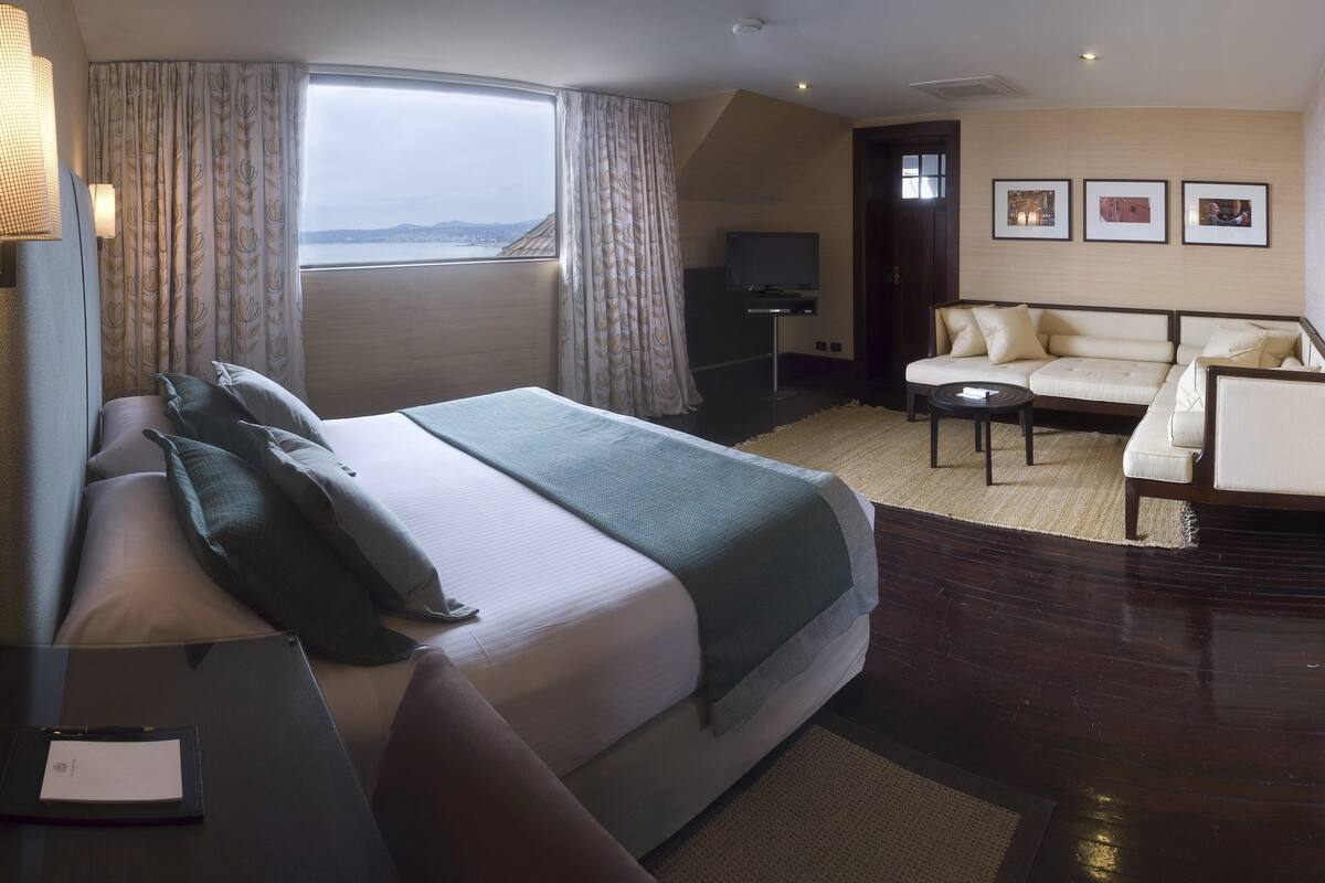 A hotel room with a bed, a sofa, a small table, and a view of the city.