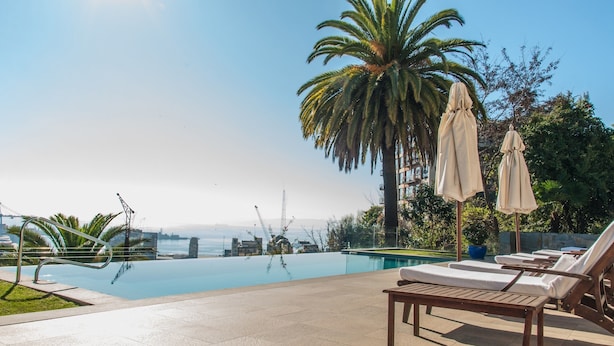Un área junto a la piscina con sillones de descanso, sombrillas y una palmera.