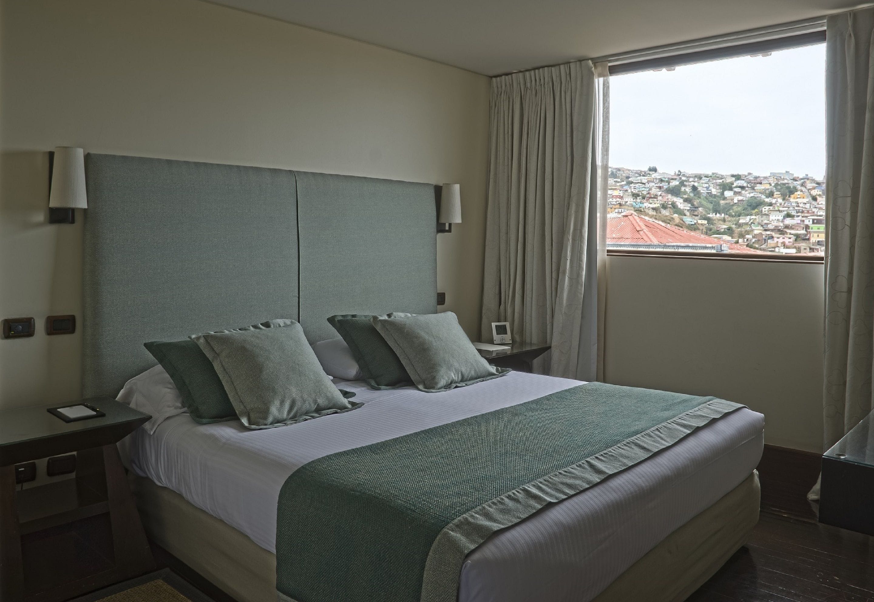 A hotel room with a large bed, two bedside tables, a nightstand, a window with curtains, and a view of a cityscape.