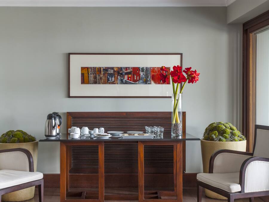 Un comedor con una mesa de madera dispuesta para dos personas, un cuadro abstracto enmarcado encima y plantas en macetas a cada lado.