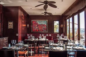 A restaurant with a high ceiling fan, a large abstract painting, and a view of the city through large windows. - Casa Higueras (Valparaiso)