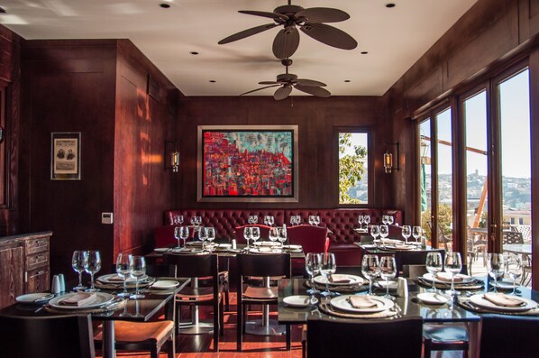 A restaurant with a high ceiling fan, a large abstract painting, and a view of the city through large windows. - Casa Higueras (Valparaiso)