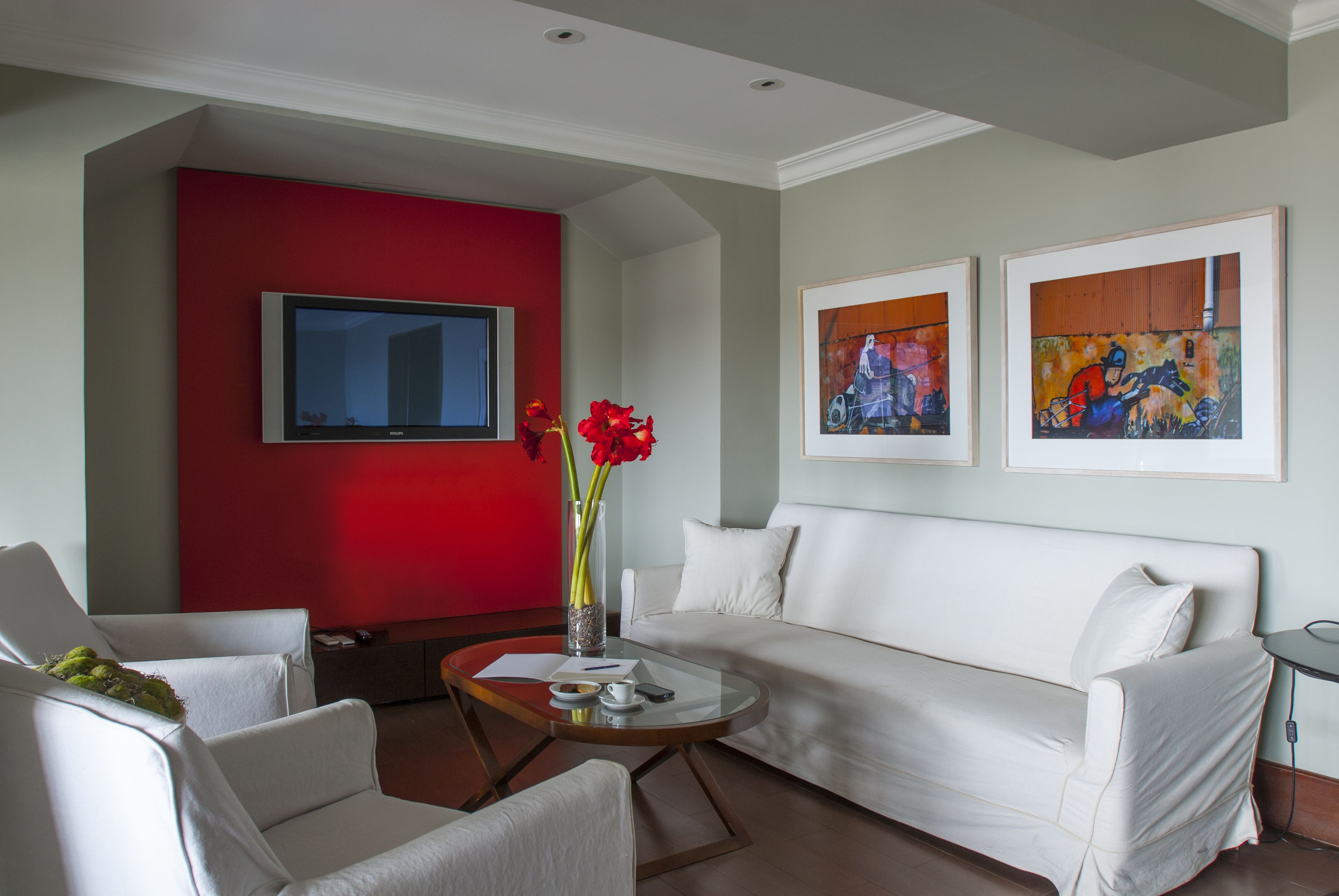 A modern living room with a white sofa, a red accent wall, a flat-screen TV, and two framed artworks.