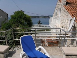 Un balcon avec une chaise bleue, un bâtiment en pierre avec un toit en tuiles et une vue sur une étendue d’eau.