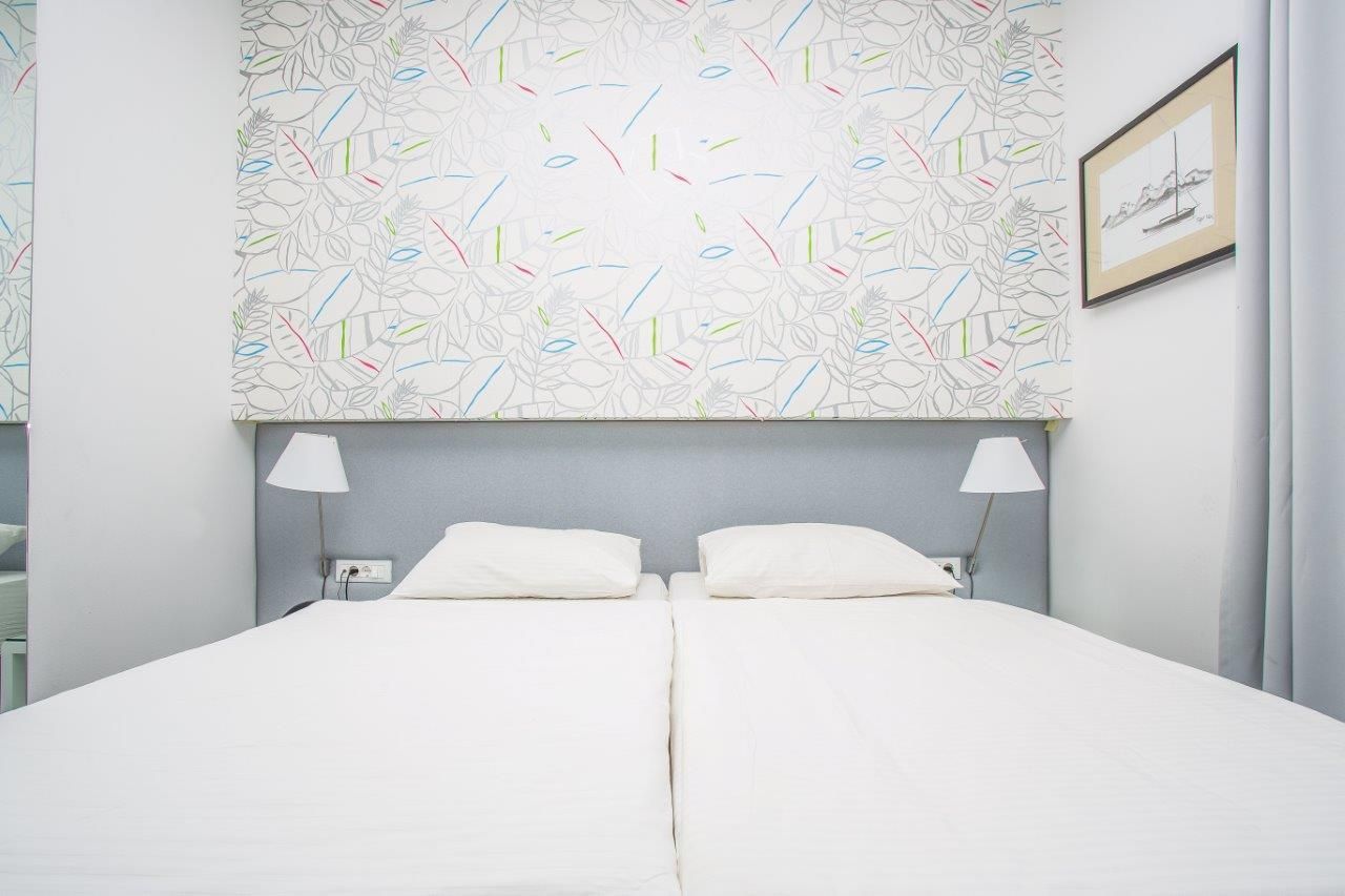 A neatly made double bed with white linens, two bedside lamps, and a patterned wall above.