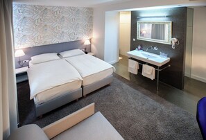 Habitación de hotel con cama doble, lámpara de noche, baño con lavamanos y espejo, y piso alfombrado.