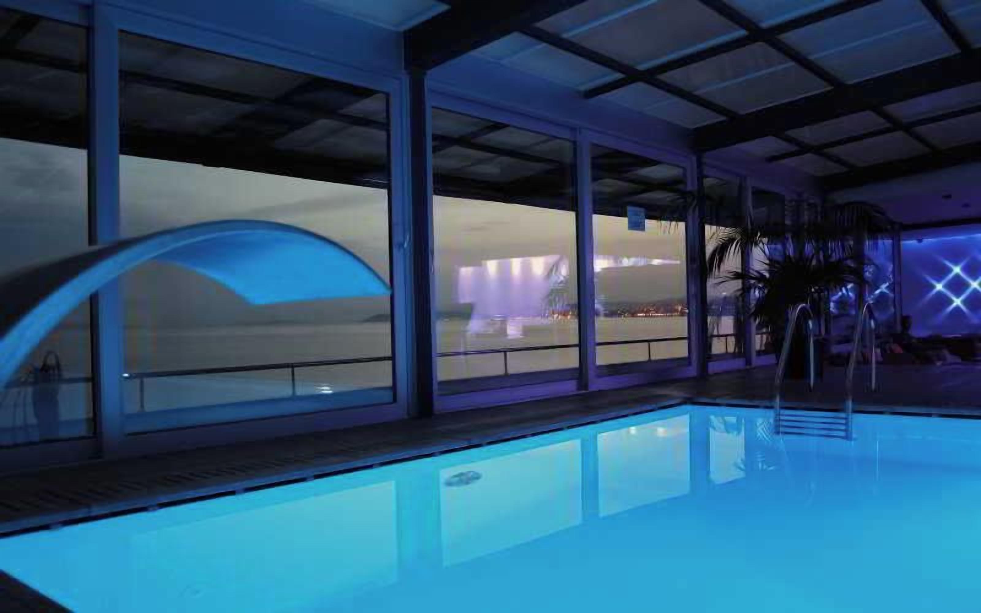 An indoor swimming pool with clear glass walls, a pool ladder, and a view of a cityscape.