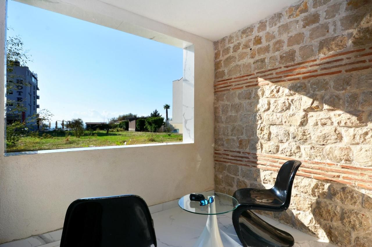 Een balkon met een stenen muur, een glazen tafel, twee stoelen en uitzicht op een gebouw en begroeiing.