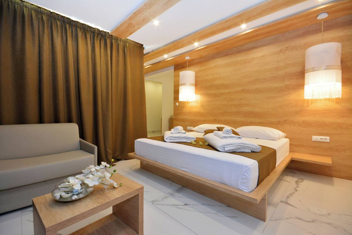 Quarto de hotel moderno com uma cama de madeira, um sofá, uma pequena mesa de madeira e um espelho grande.