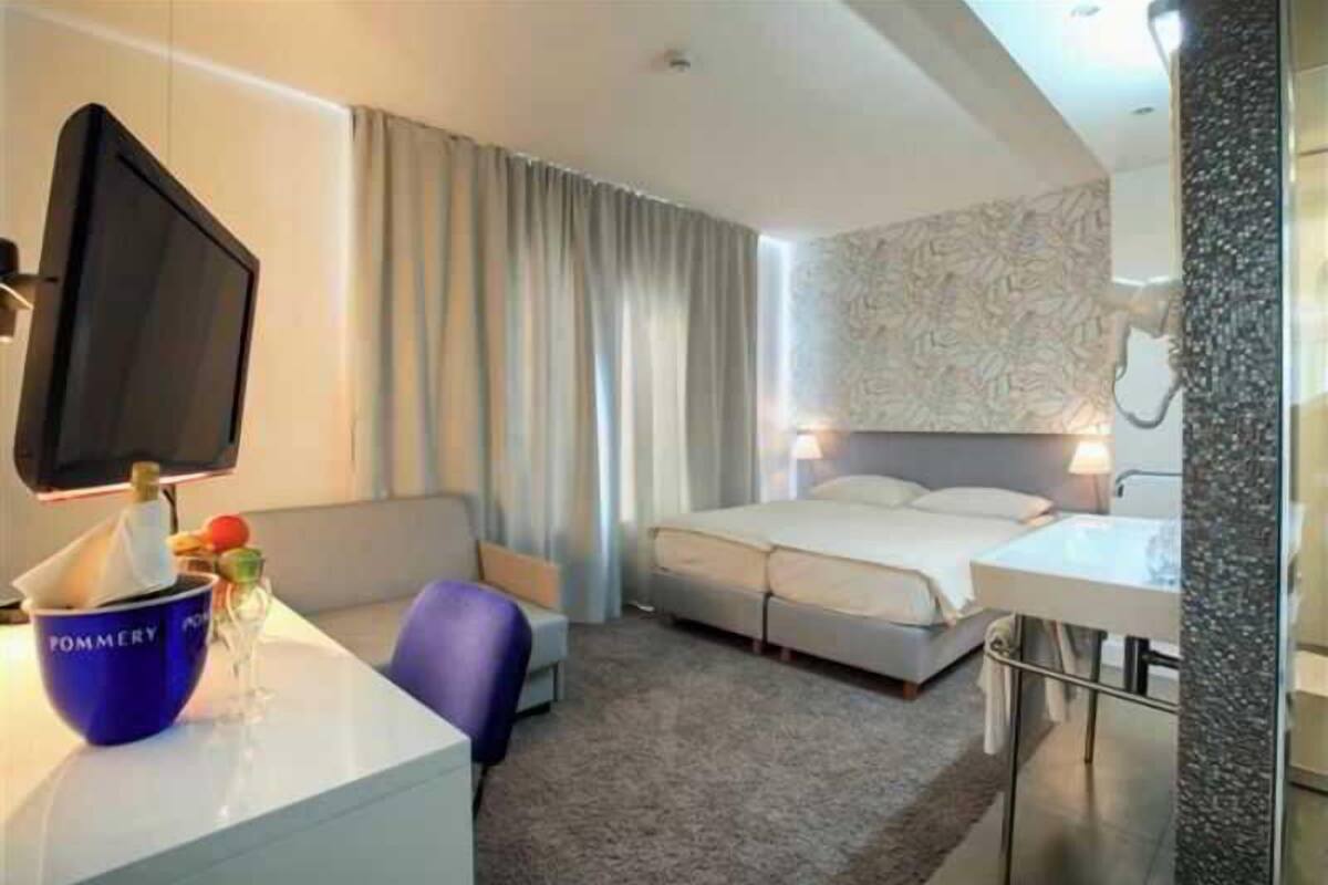 Quarto de hotel com cama, escrivaninha com computador, cadeira, televisão e banheiro com pia e espelho.