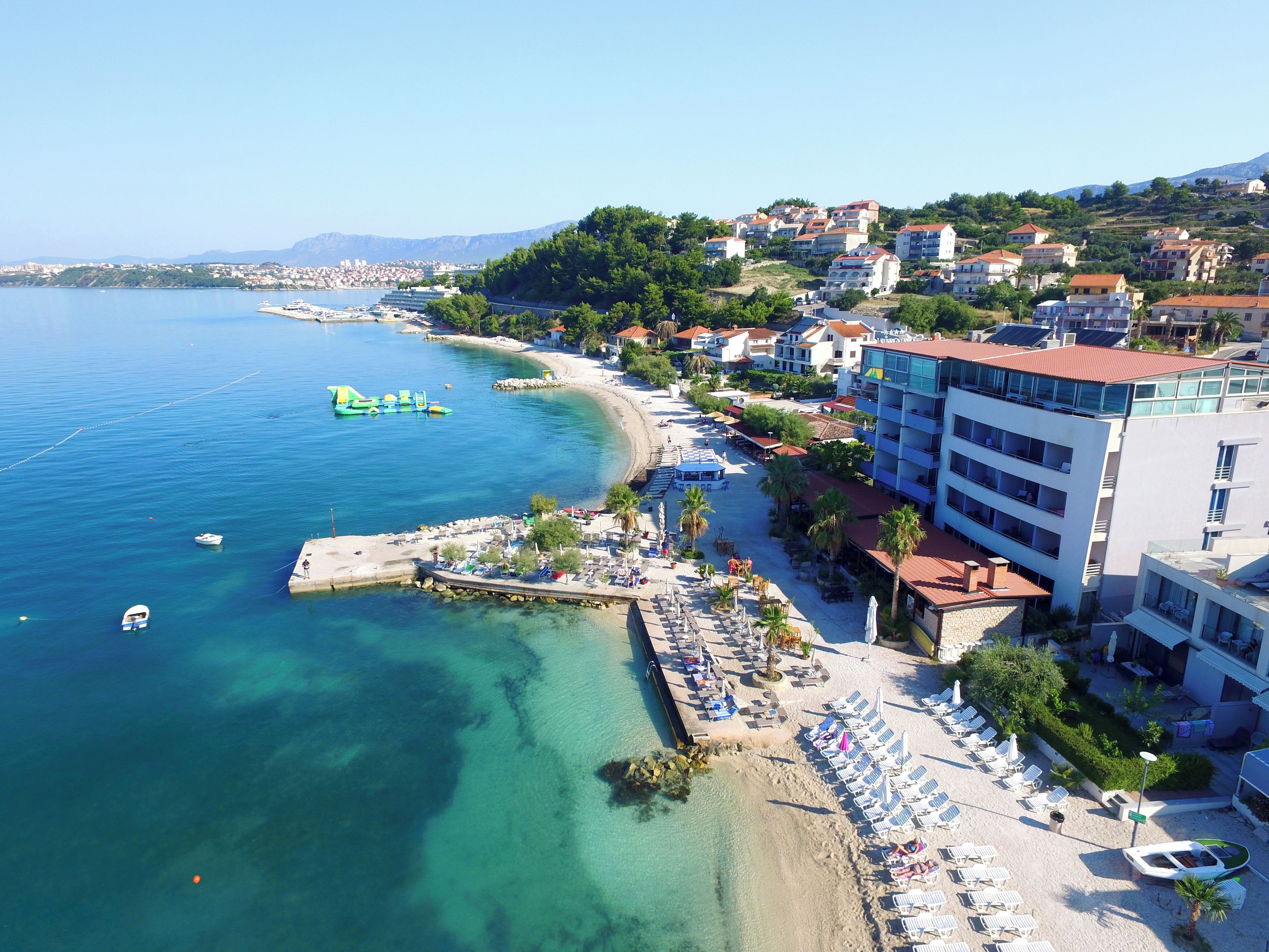 Un hotel frente al mar, con playa, aguas cristalinas y vistas a la ciudad al fondo.